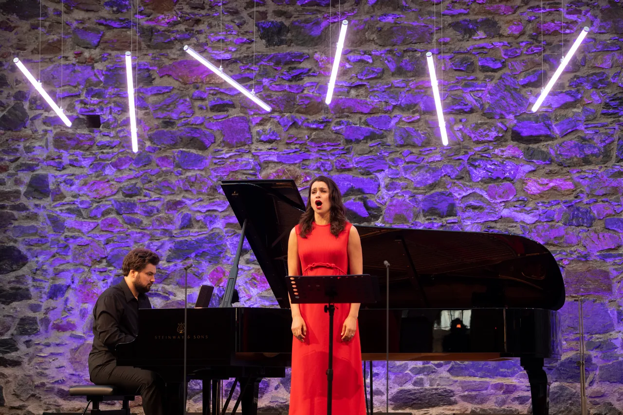 Asmik Grigorian canta con Lukas Geniušas al piano, ante el muro de piedra iluminado en violeta del castillo de Taggenbrunn