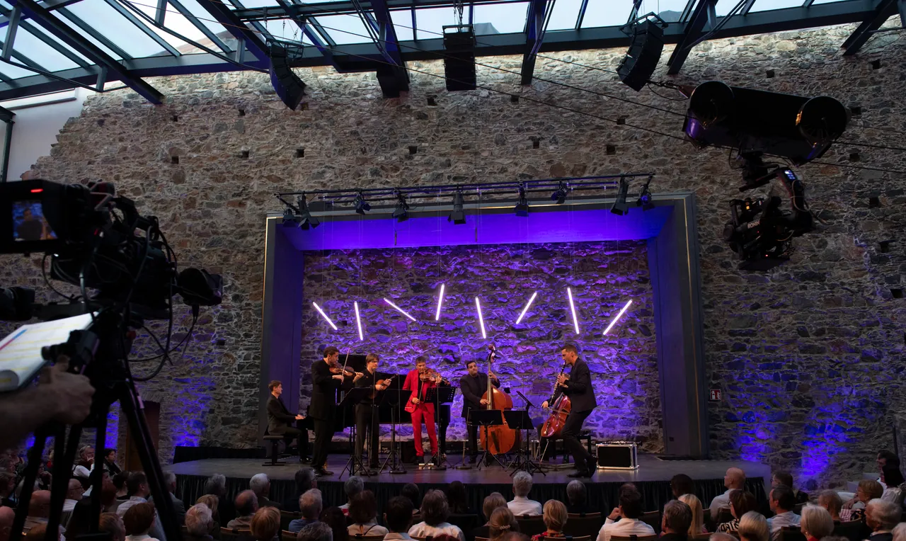 Una cámara de televisión en primer plano; al fondo, Philharmonix en el escenario del castillo de Taggenbrunn y, arriba, la cámara por cable